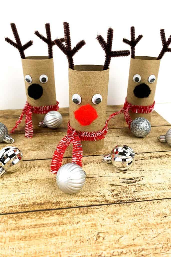 vertical image of a set of three reindeer paper roll crafts with pipe cleaner antlers and pom pom noses set on a holiday wood background