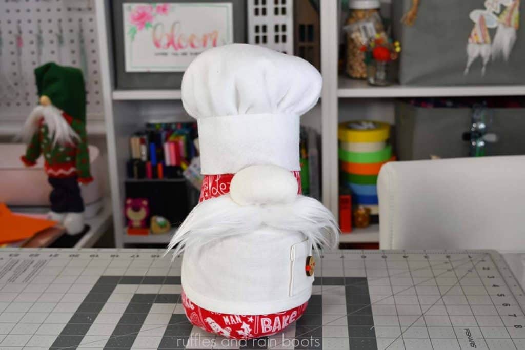 a Christmas gnome with white and red holiday fabric body, white chef apron pattern, and white chef hat with white mustache and large nose in front of a craft room background