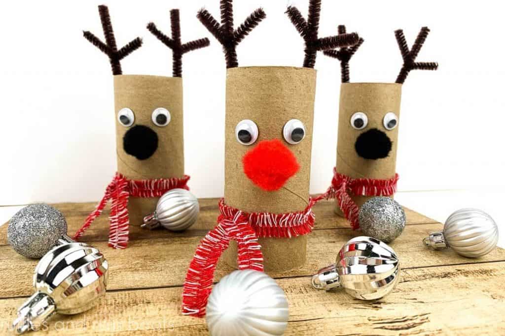 close up image of three reindeer paper roll crafts on a wood base and white background with silver Christmas ornaments scattered around