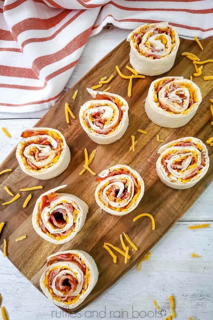 eight bacon ranch pinwheels served on a wood cutting board with shredded cheese scattered and a red and white French stripe dish towel on a white wood background