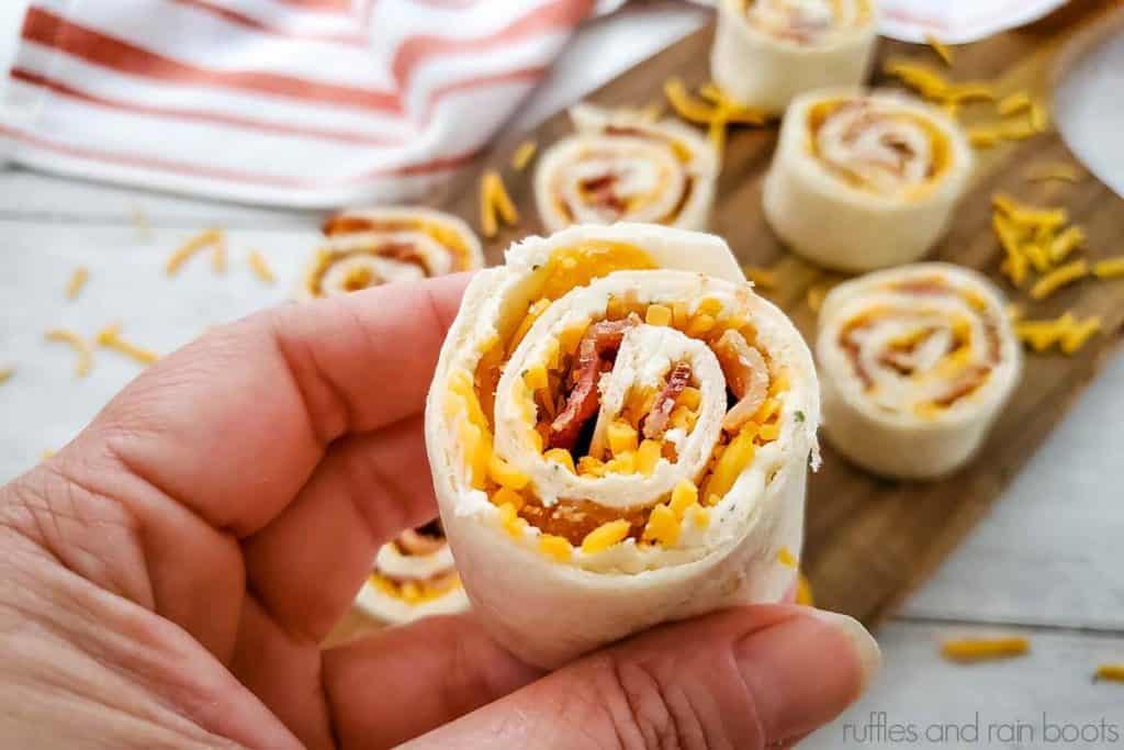 close up of a hand holding a bacon ranch pinwheel made with a tortilla over a cutting board of pinwheels and a dish towel