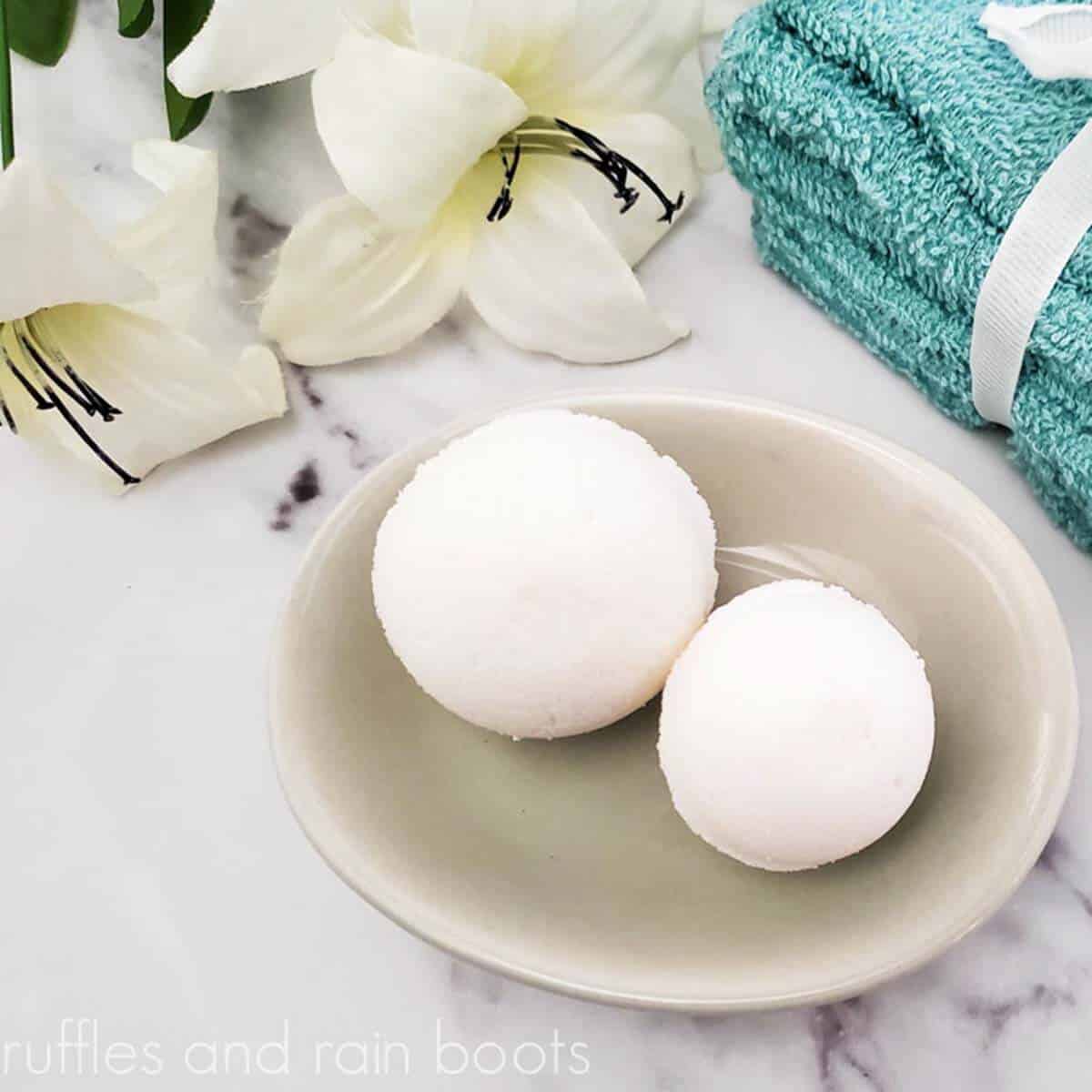 Square image close up of 2 sizes of homemade milk bath bombs on a soap dish with a vanilla blossom and teal washcloths all on a marble counter top. 