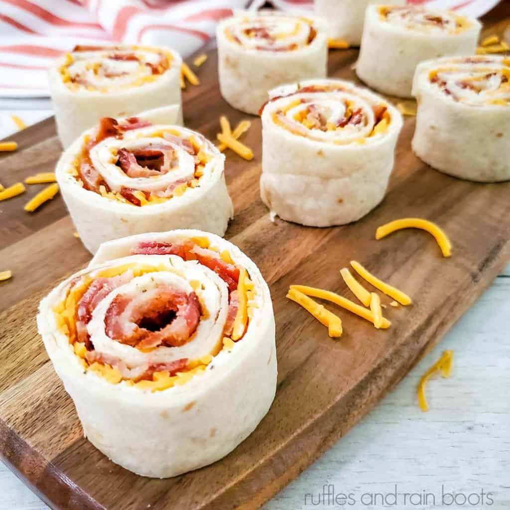 square image of a close up of a bacon ranch pinwheel on a wood cutting board with shredded cheese and a french stripe dish towel