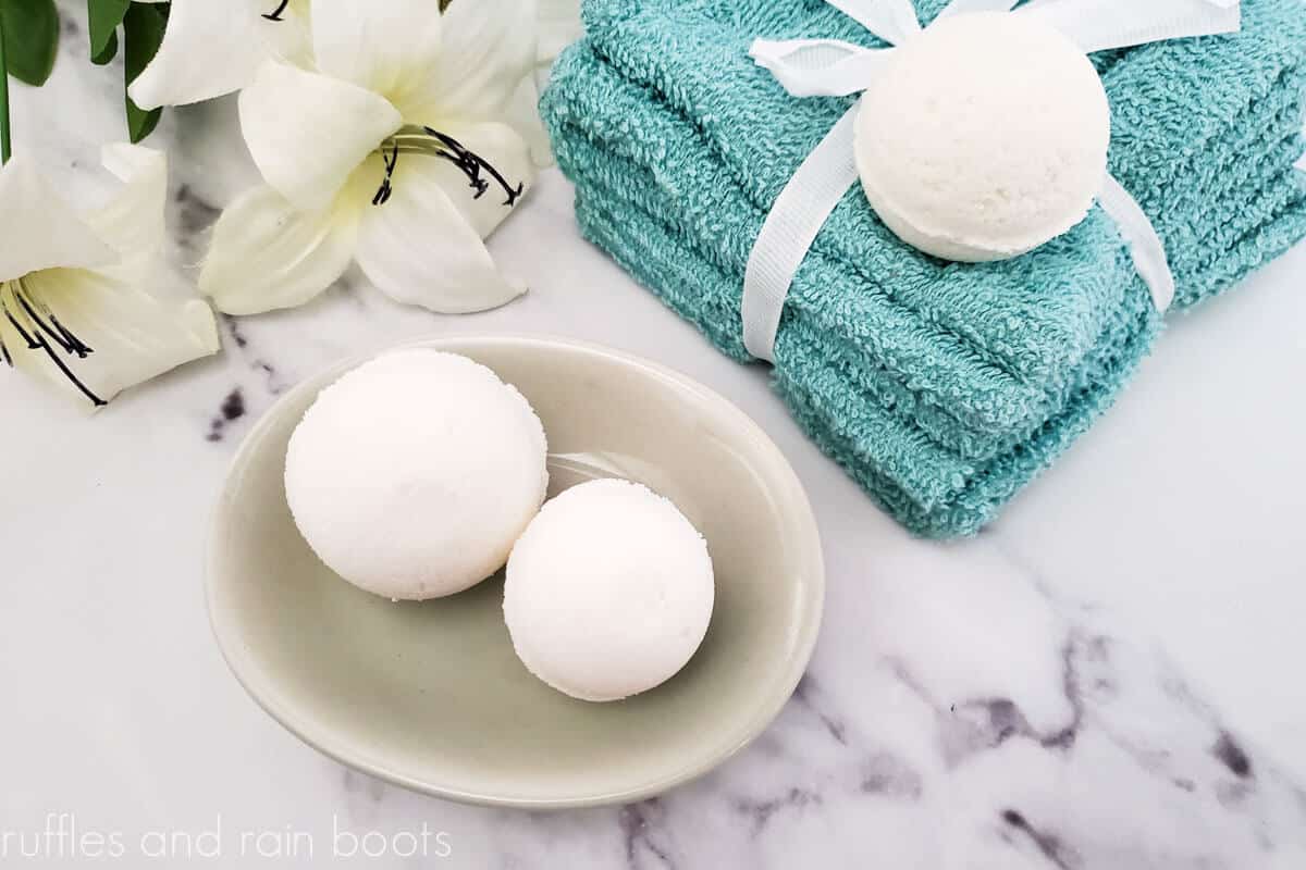 Horizontal image showing three cream milk bath bombs in a soap dish and on a teal washcloth stack on a white and gray marble counter top with vanilla blossoms.