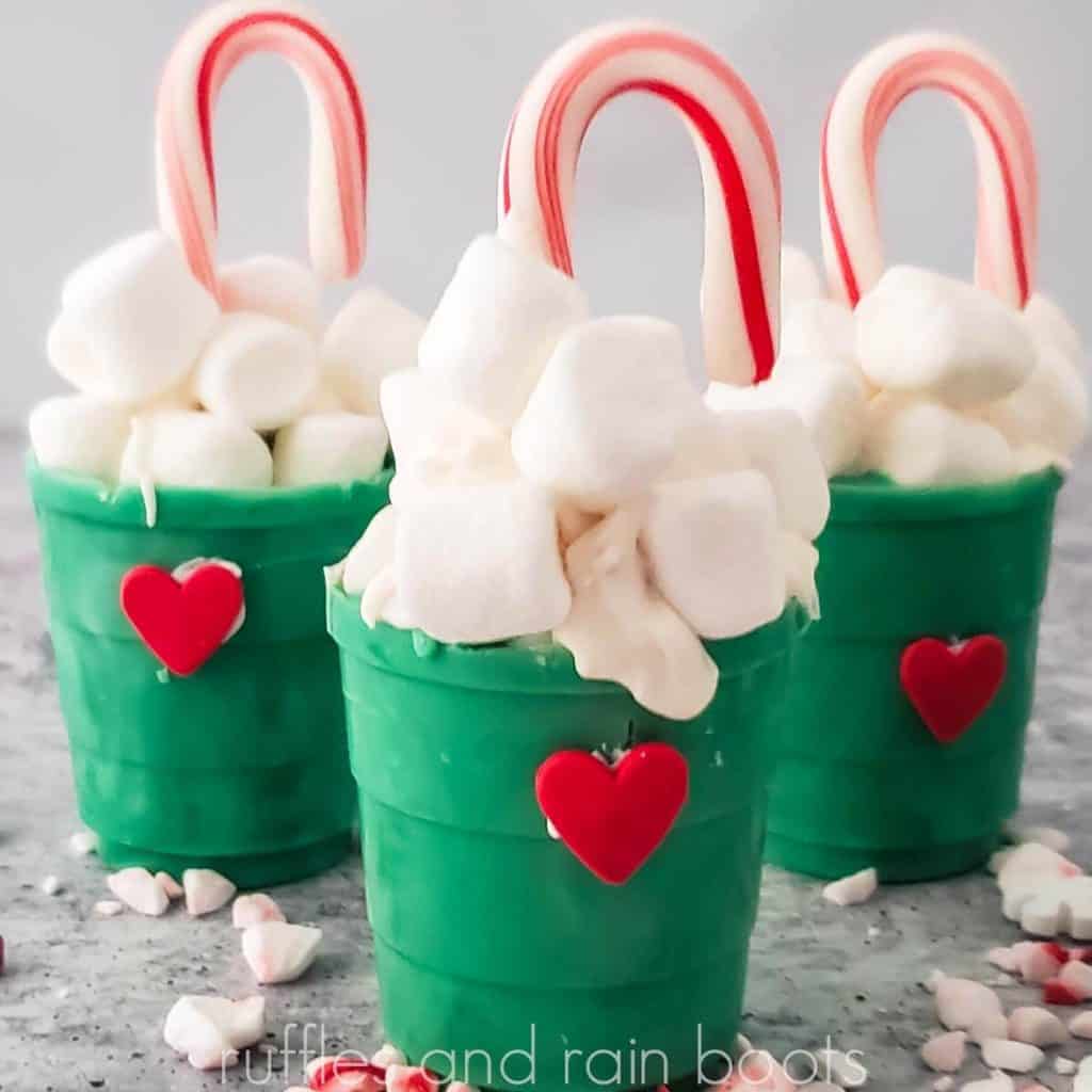 square image of three green chocolate candy cups in a Grinch theme with marshmallows and mini peppermints on a gray and white background.