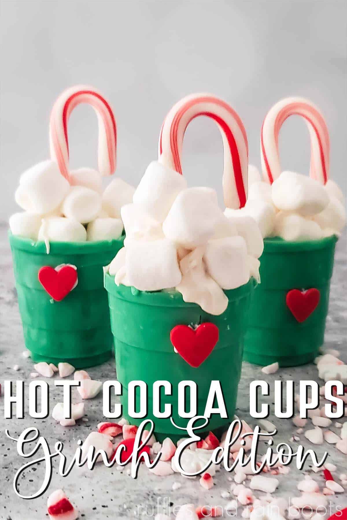 vertical close up image of gray background with three green chocolate cups with red candy heart, mini marshmallows, and candy canes with text which reads hot cocoa cups Grinch Edition ruffles and rain boots.