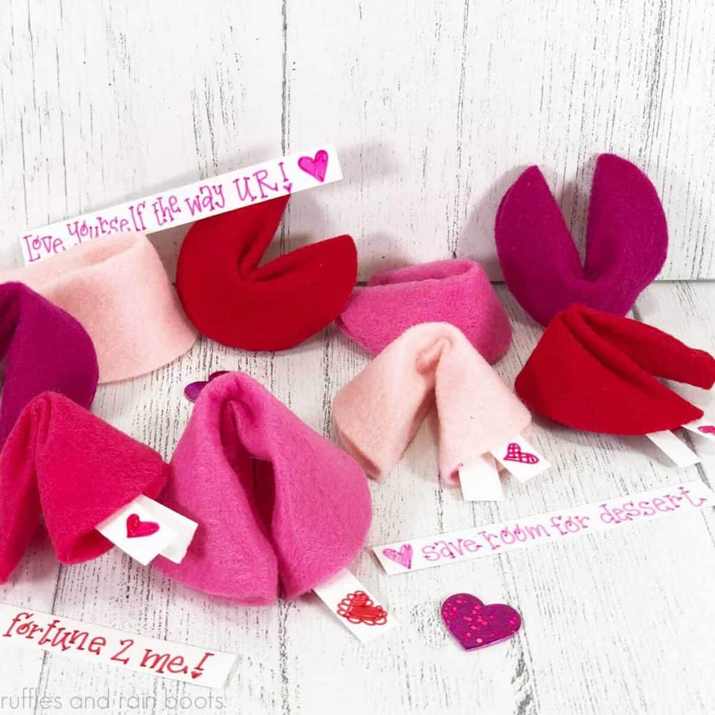 square image of pink and red felt fortune cookies made for Valentine's Day placed on a white wood background with pink glitter heart confetti