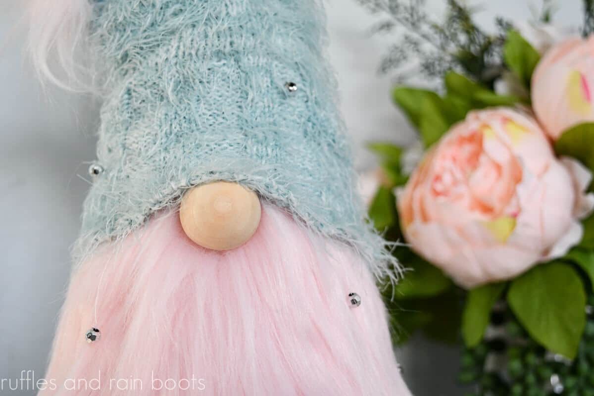 Horizontal close up image of a cone gnome made with pink faux fur, wooden ball for a nose, and a sweater sleeve for a hat in front of a vase with pale pink peonies.
