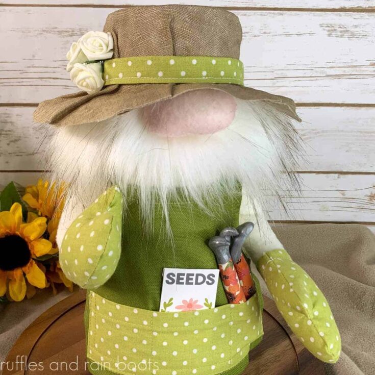 close up square image of the gardening gnome pattern from ruffles and rain boots styled in a linen sun hat with flowers and green canvas apron with seed packet and tools