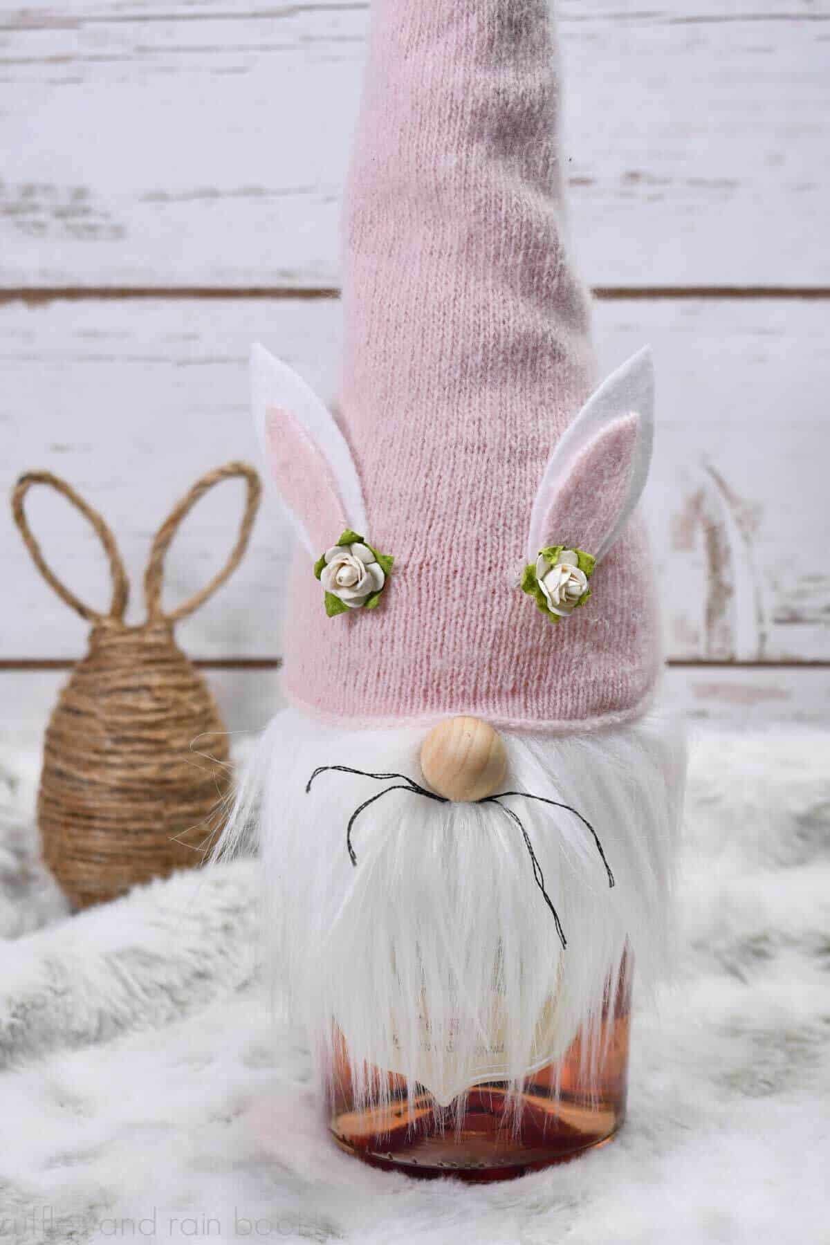 Vertical image of a pink hat Easter gnome bottle topper with ears, whiskers, and beard in front of a twine bunny egg on a fur table with wood slat background.