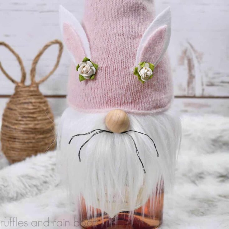 Pink hat Easter gnome bottle topper DIY with white beard and whiskers on a white fur background in front of a wood wall.