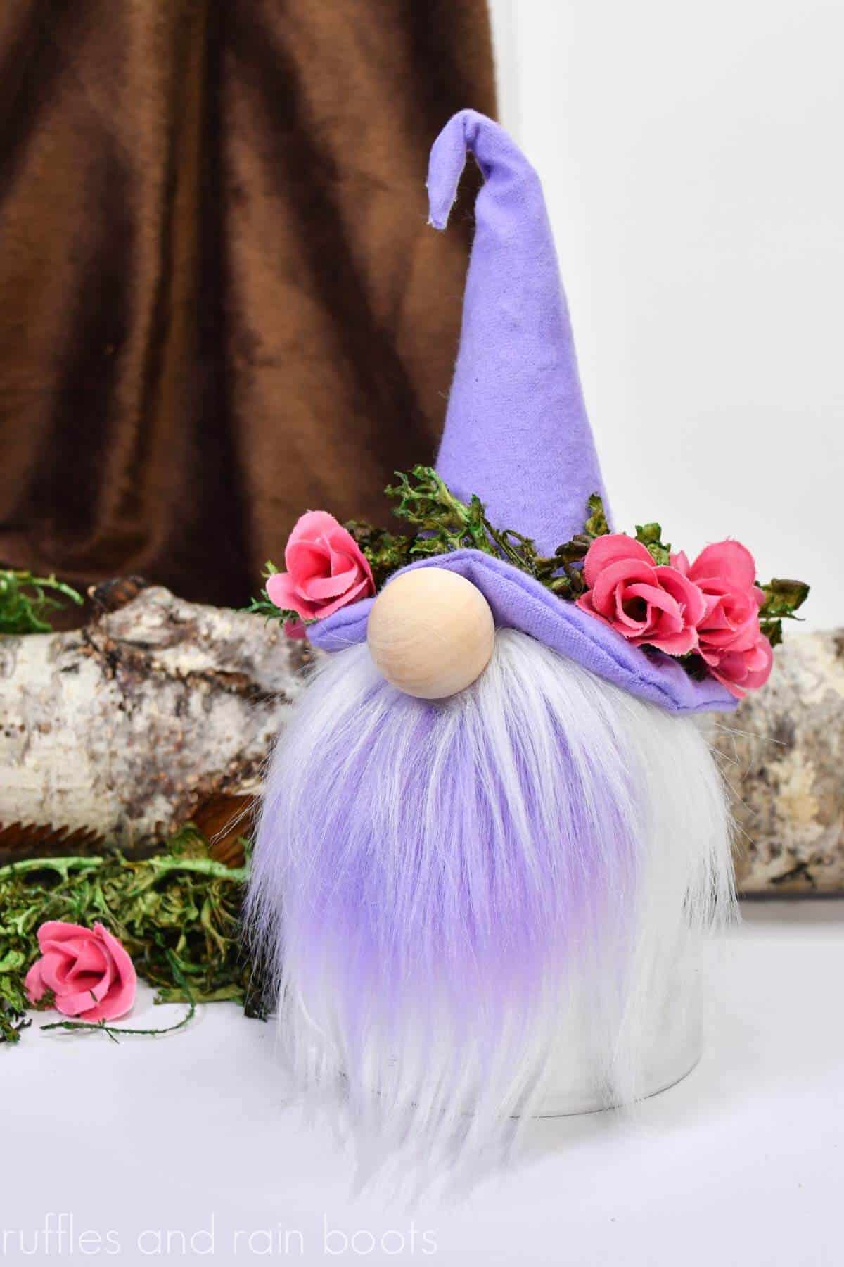 Vertical image of a Spring clay pot gnome made with wizard hat pattern, floral crown, and heart shaped two color gnome beard in front of a brown and white background with log, moss, and pink flowers.