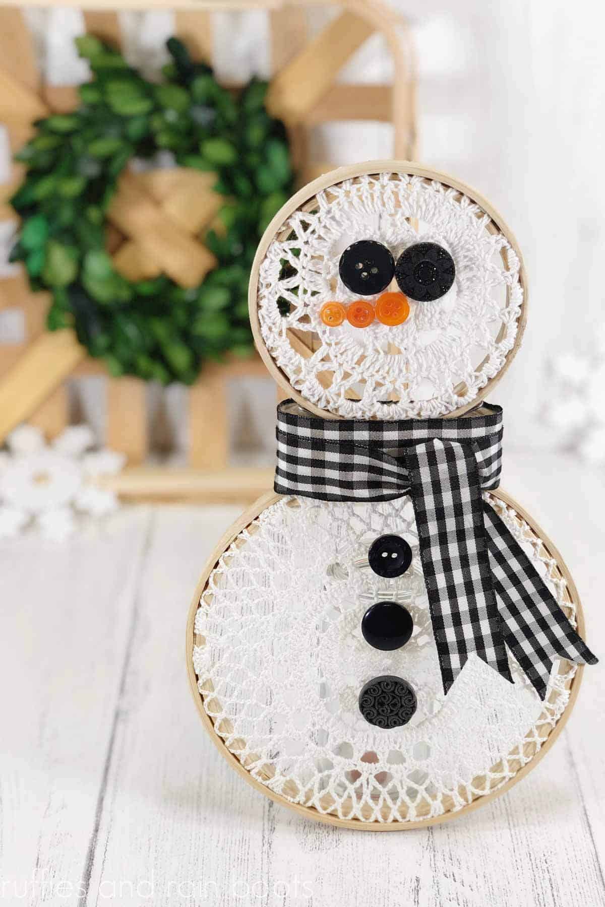 Vertical image showing a white lace snowman made from small embroidery hoops and pre-made cotton doilies in front of a farmhouse background of green wreath, tobacco basket, and white wood.