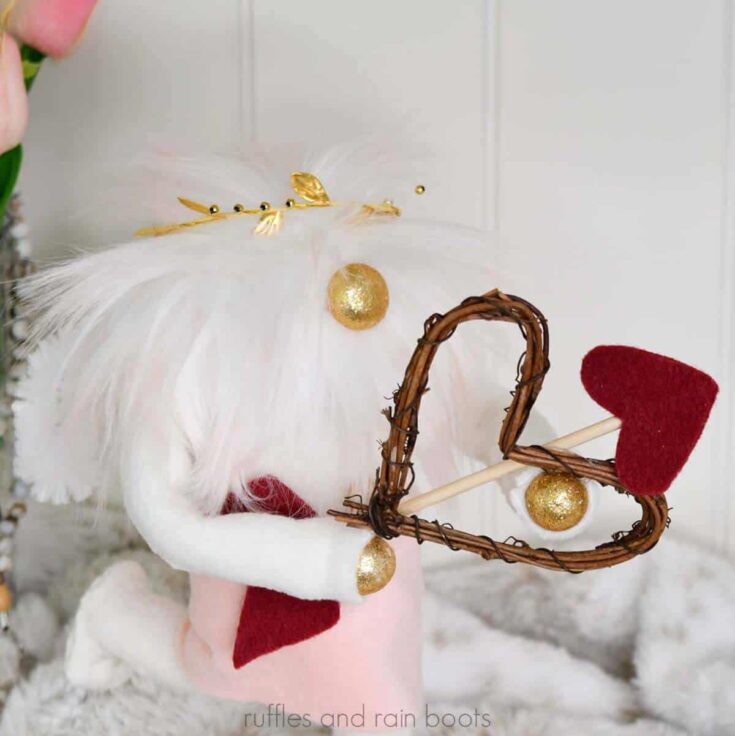 Square close up image of a white and pink haired Cupid gnome DIY holding a bow and arrow from hearts with one leg kicked back standing in front of fa fur background with tulips.