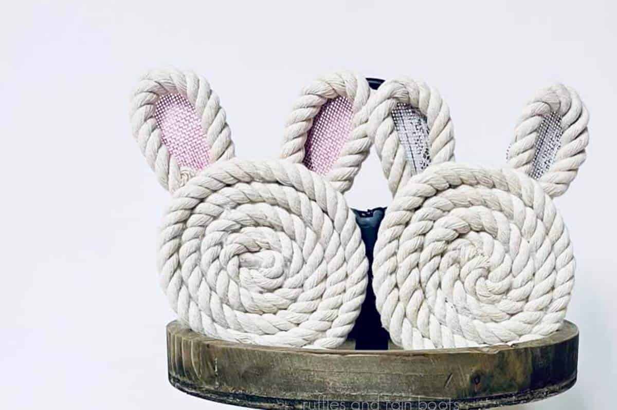 Two bunny coasters made with Dollar Tree white rope and ribbon placed on the top shelf of a wood tiered tray and placed in front of a light gray wall.