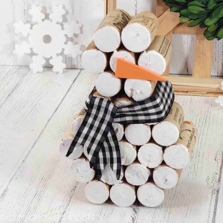Close up, off center image of a cork snowman with a Buffalo check ribbon scarf, an orange carrot nose made from foam and cork ends painted white in front of a farmhouse background with a snowflake and white wood.