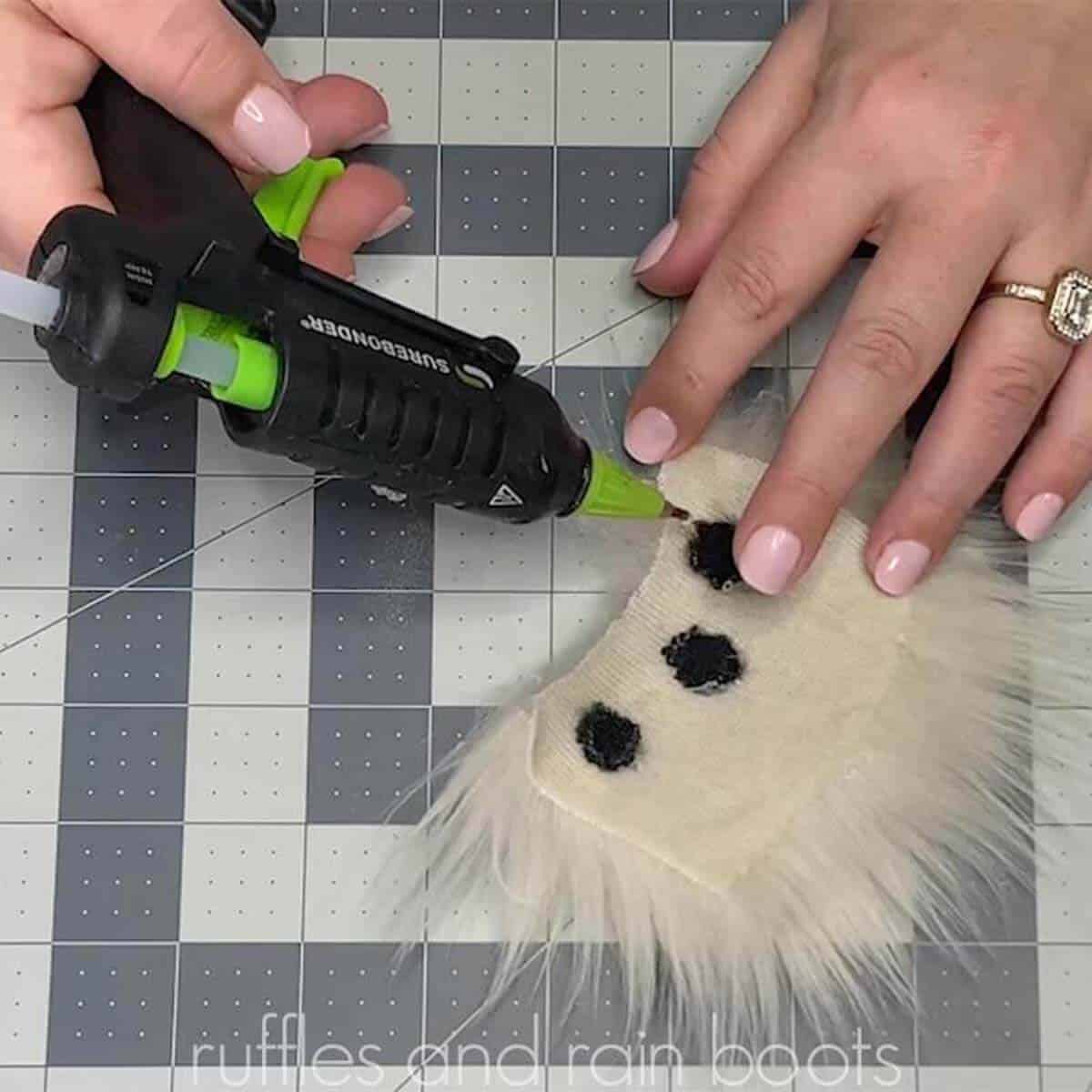 Square close up image of a glue gun attaching black fur insert pieces into a blonde fur block to create a custom gnome beard.