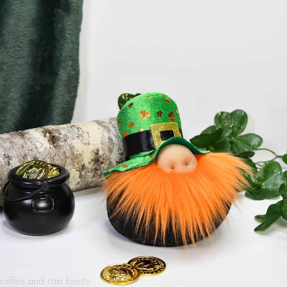 Close up square image showing a DIY leprechaun gnome for St Patrick's Day with green and gold hat, orange fur beard, and round black body sitting on a white table in front of green clover and a pot of gold coins.