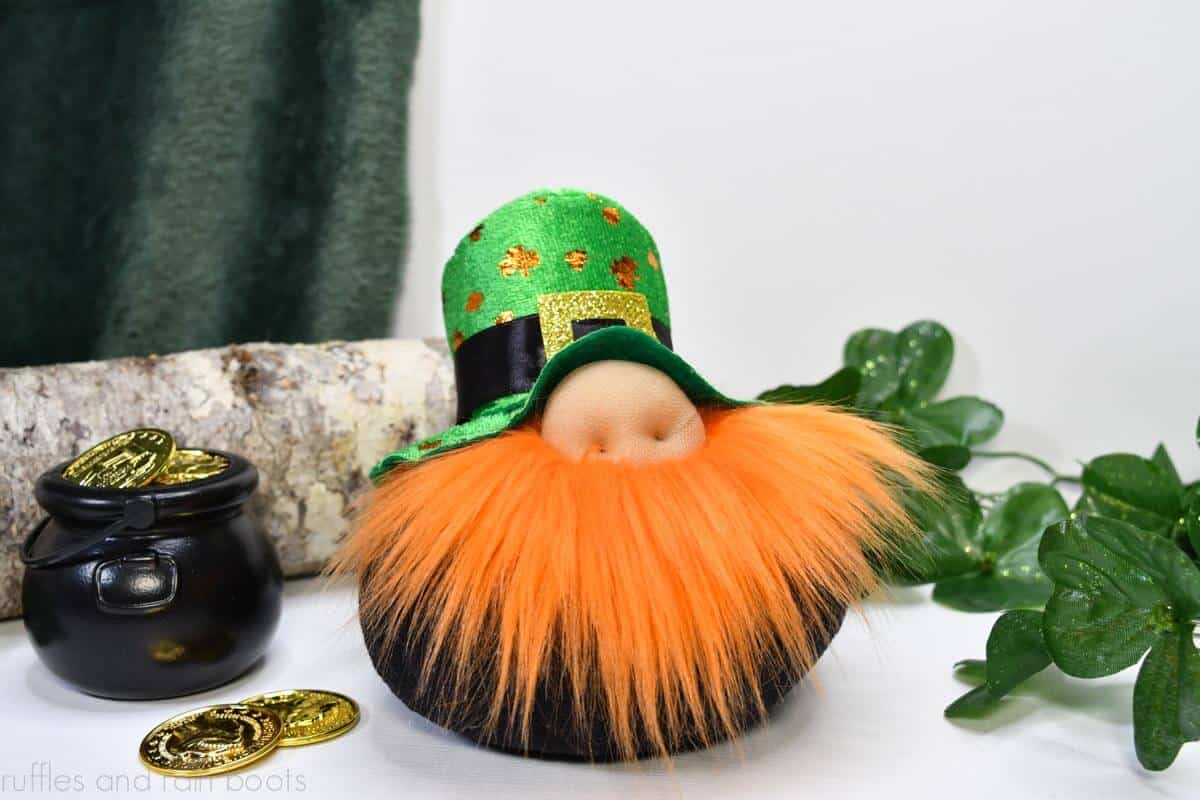 Horizontal close up image of a leprechaun St Patrick's Day gnome with a Dollar Tree hat, wrap around beard in orange fur, and a round body in front of a white and green background with clover, coins, and a pot of gold.