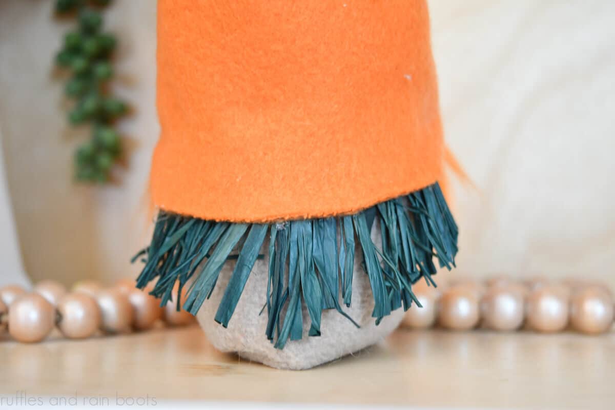 Horizontal close up image of a carrot gnome DIY project made with fleece and green raffia for Easter decor in front of a farmhouse bead garland and succulent..