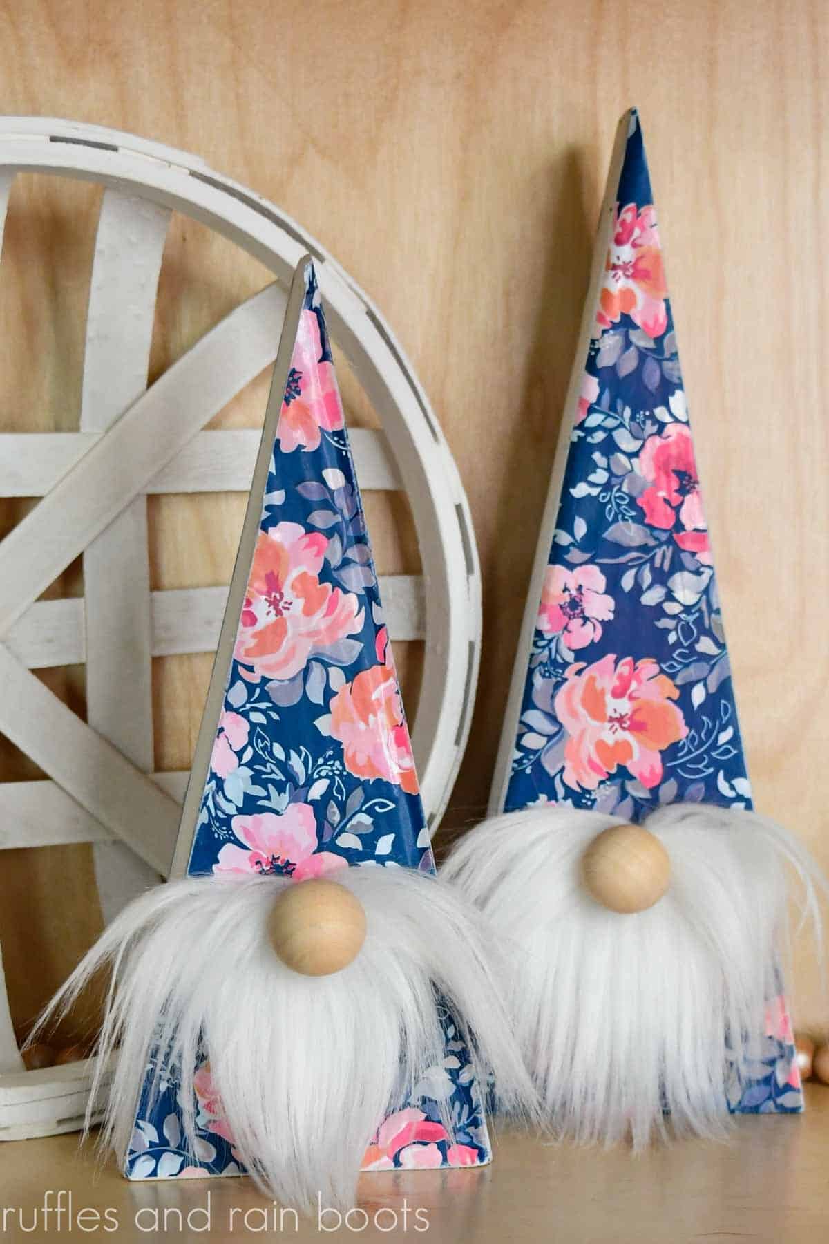 Vertical image of a pair of Spring wood gnomes with colorful floral vinyl from the Dollar Tree and white beards in front of a light wood background and a white tobacco basket.