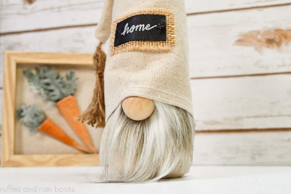 Horizontal image of a close up of a farmhouse gnome made with drop cloth, faux fur, a tassel, and a burlap and leather patch in front of a white wood wall and a carrot sign.