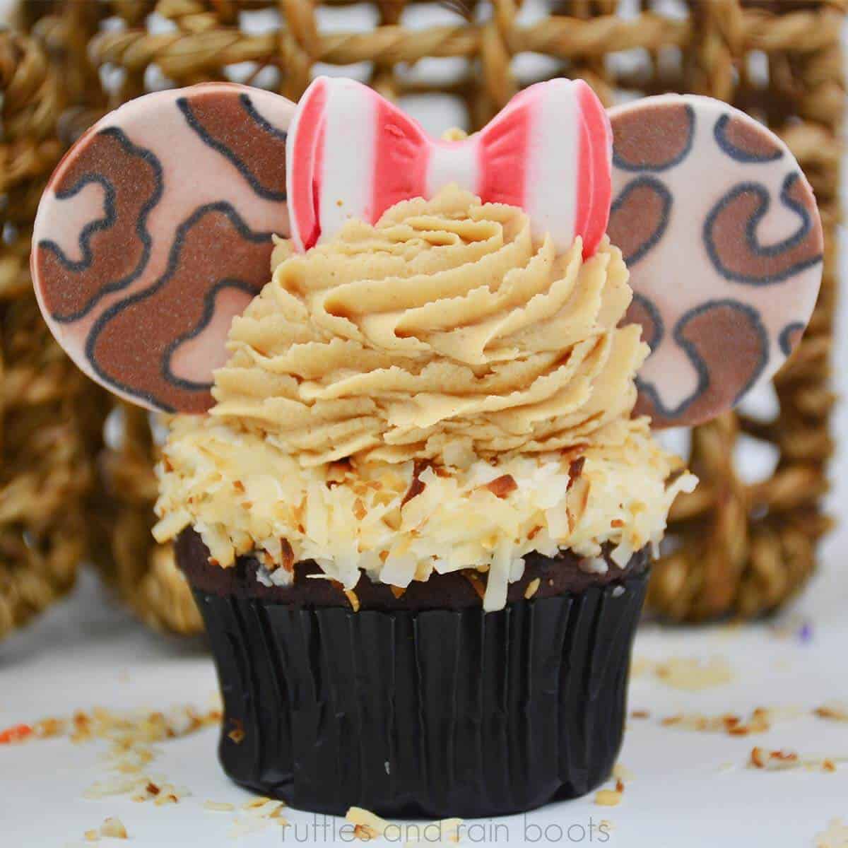 Square close up image of a safari Minnie ear cupcake with a stripe red and white bow in fondant with peanut butter frosting and coconut topped chocolate cupcake in front of a wicker basket.