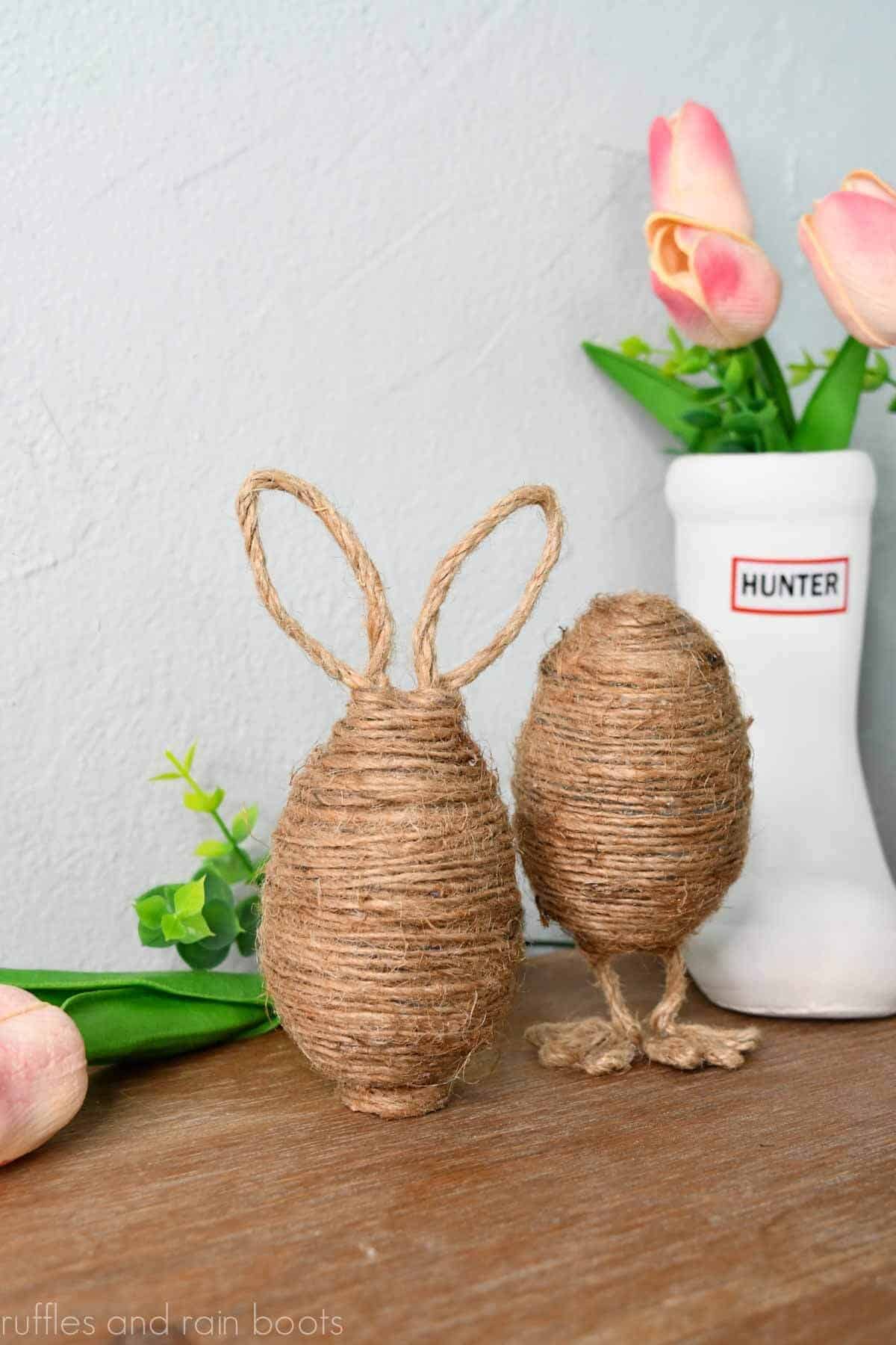 Vertical image with twine wrapped plastic eggs on a wood table in front of a light gray wall with tulips and spring greenery.