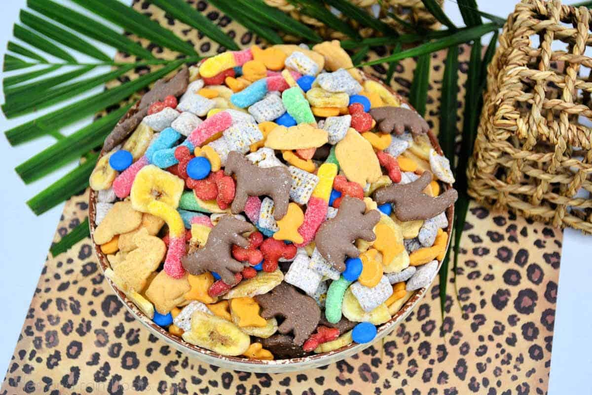 Horizontal image showing a cheetah print background, large green leaf, and a big, colorful bowl of Jungle Cruise snack mix for a safari party, Wild One birthday, or movie night.