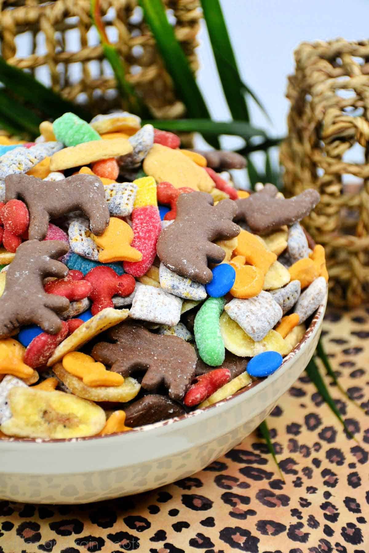 Vertical image of a cheetah print background with wicker baskets and leaves showing a colorful, fun safari Jungle Cruise snack mix treat in a white bowl.