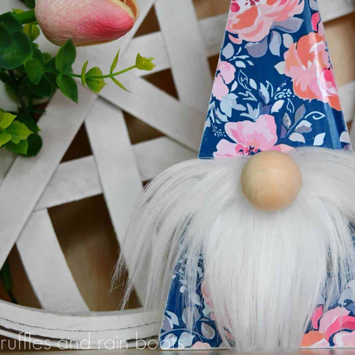 Square close up image of a triangle Spring wood gnome with white fur and wood nose in front of a tulip and white tobacco basket.