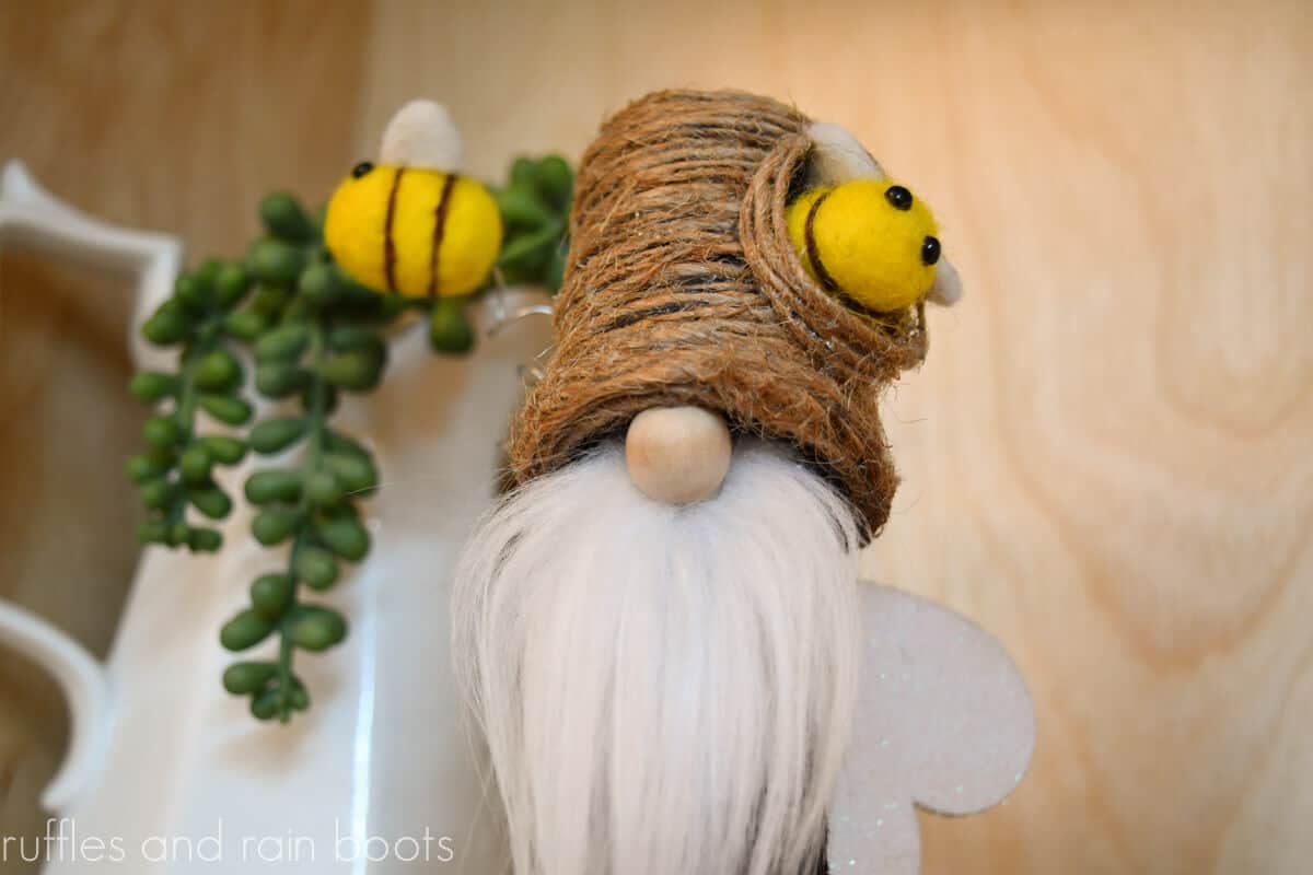 Horizontal close up image of a mini bee gnome with a beehive hat and flying felted bees, a wood ball nose, and a white faux fur beard.