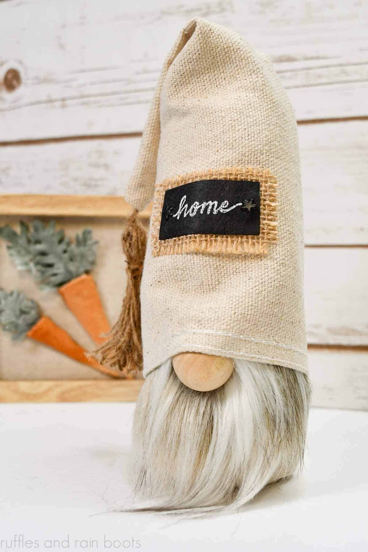 Vertical image of a simple, rustic gnome made with a canvas fabric with a free gnome pattern, faux fur, and a wood bead nose in front of wood background and tiered tray carrot sign.