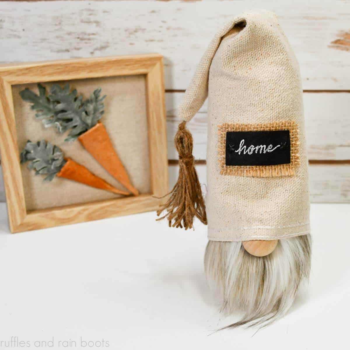 Square image close up showing a top down look of a drop cloth gnome in a farmhouse style with tassel and leather and burlap patch in front of a wood wall and a carrot shelf sitter sign.