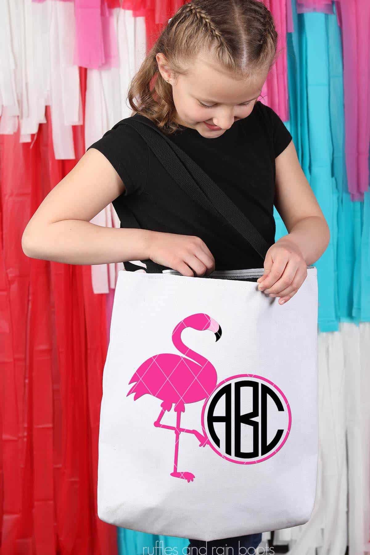Vertical image of a pink flamingo monogram SVG on a canvas tote bag on the shoulder of a young girl in a black t shirt standing in front of a red and teal party back drop.