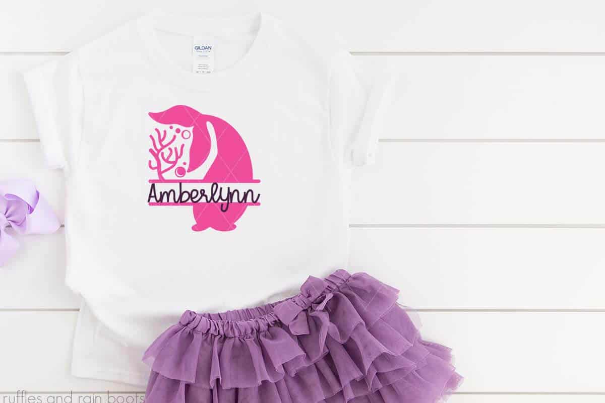 Horizontal image of little girl white t shirt with pink vinyl mermaid tail monogram SVG with Amberlynn name with a purple tulle skirt and hair bow on white wood background.