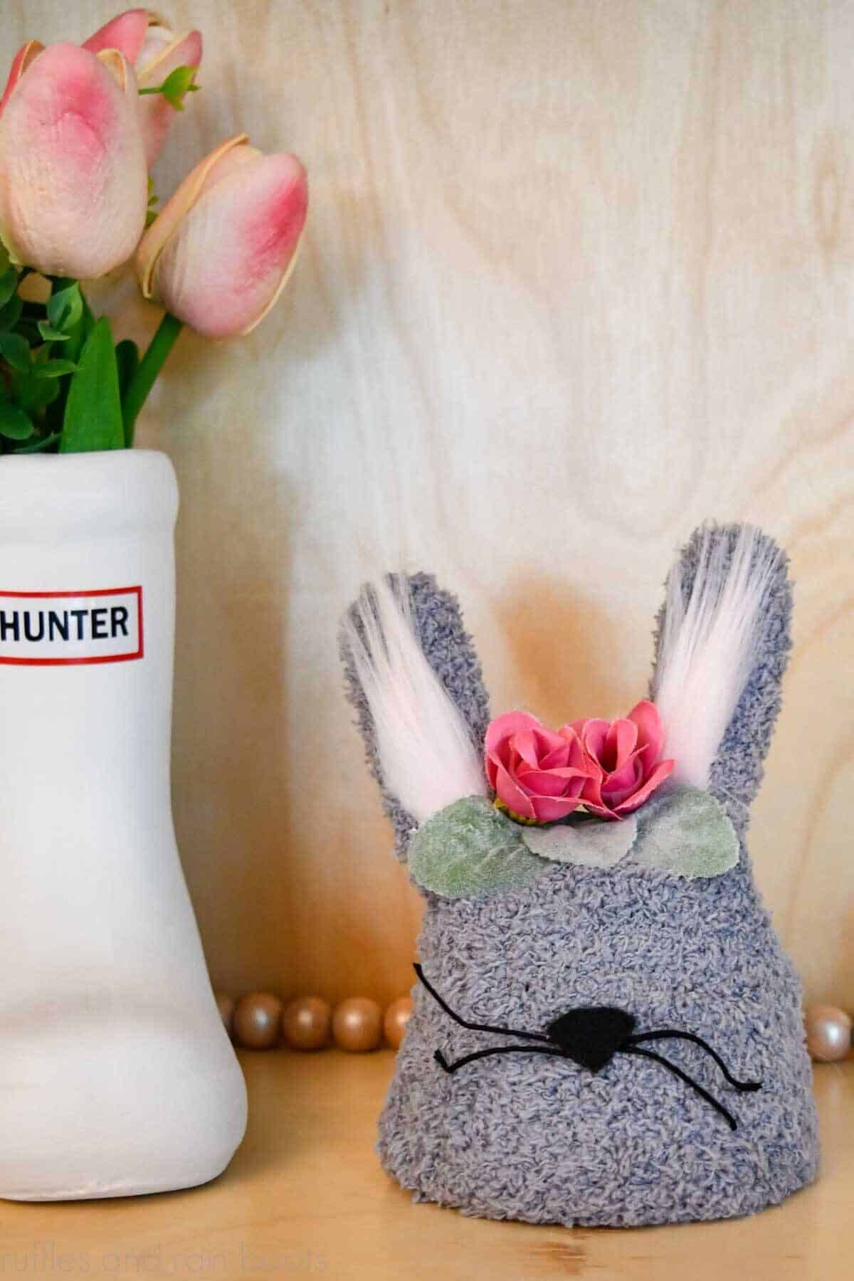 Vertical image of a gray tiered tray bunny craft made from a glove and clay pot standing in front of a wood background next to a white rain boot planter with tulips.