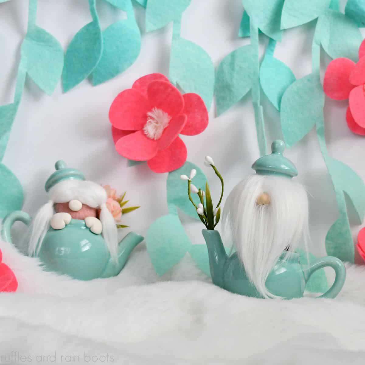 Square close up image of a boy and girl gnome in teal teapots on fur in front of a white wall with felt hanging vines and pink flowers.