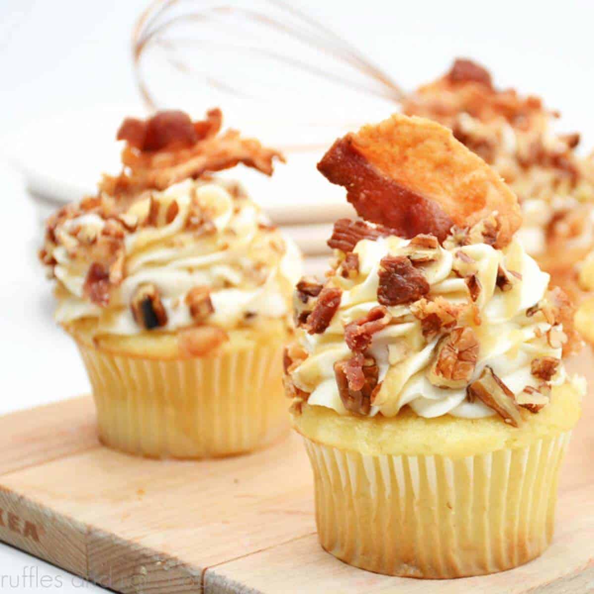 Close up square image of three maple bacon cupcakes on a small wood cutting board in front of a whisk.