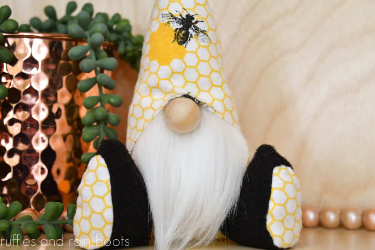 Horizontal image close up of a mini gnome for tiered trays with a bee and hexagon fabric hat sitting next to farmhouse beads and a copper vase with succulent.