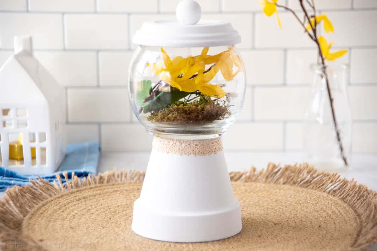 Horizontal image of white terrarium made of a clay pot, saucer, wood bead, and a fish bowl vase from the Dollar Tree set on a jute placement in front of a white tile wall.