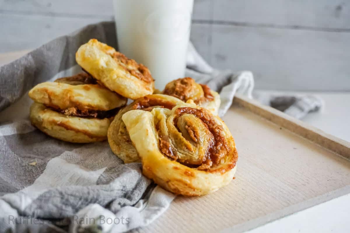 Horizontal image of peanut butter and jelly pinwheel sandwiches stacked on a wood tray with a kitchen towel and glass of milk.