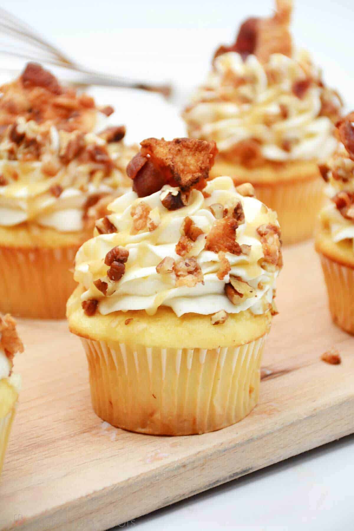 Vertical image of a close up of a whiskey maple bacon cupcake recipe topped with chopped pecans, bacon, and a whiskey caramel drizzle sauce.