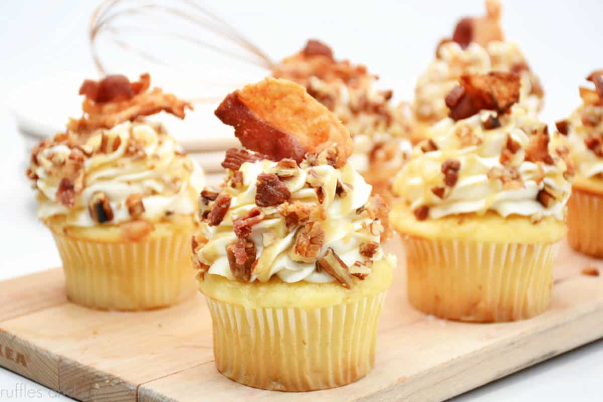 Close up horizontal image of maple bourbon bacon and pecan cupcakes topped with whiskey sauce, crushed pecans, and chopped bacon pieces styled on a small wood cutting board on a white background.