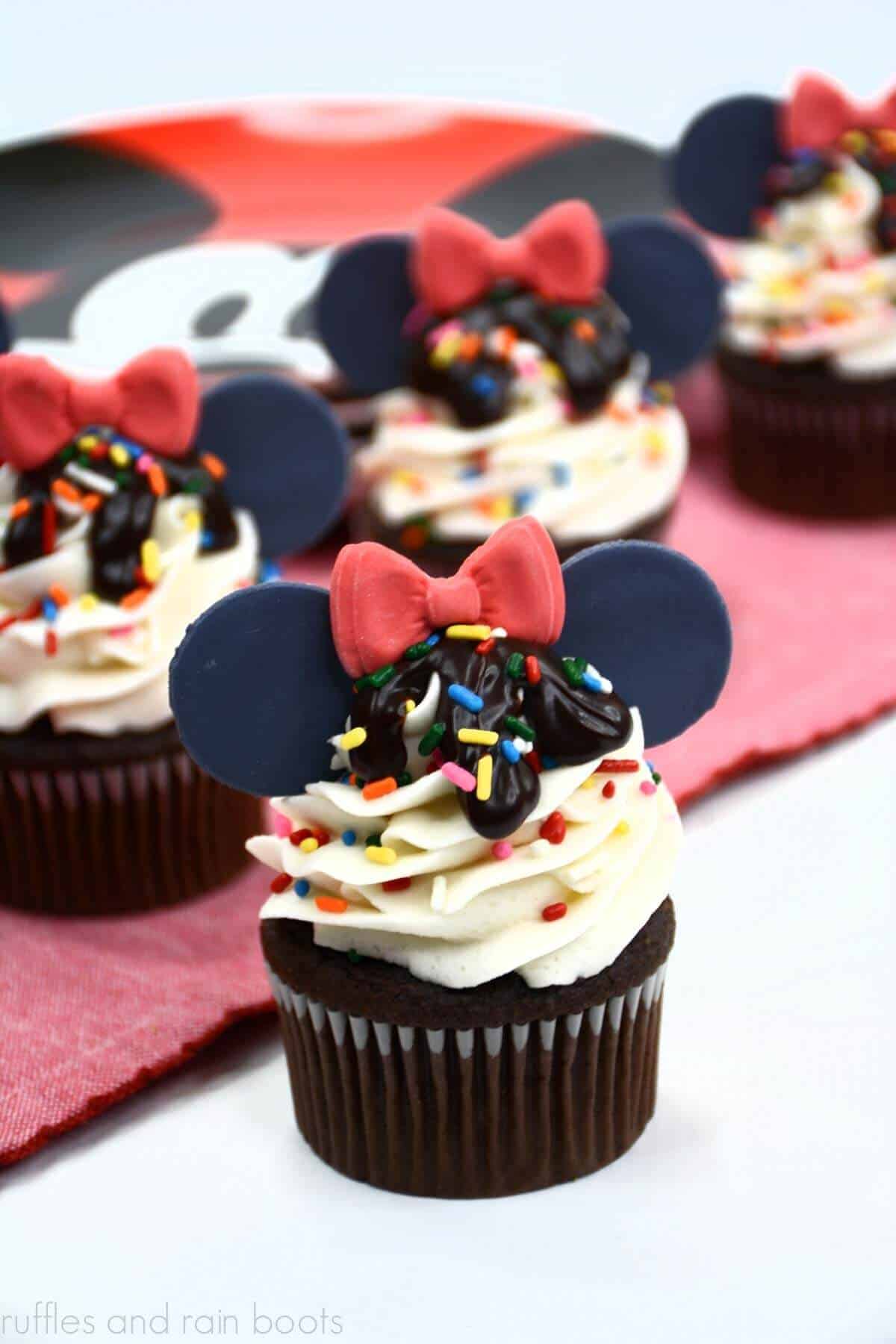 Vertical image of four ice cream sundae cupcakes decorated with Minnie Mouse bow and fondant ears on red Mickey Mouse background.