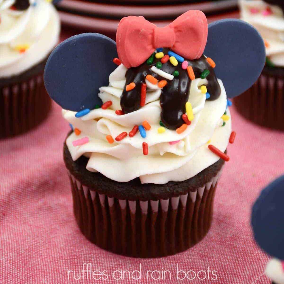 Square close up image of a Minnie Mouse ear and bow ice cream sundae cupcake with vanilla frosting, sprinkles, and easy chocolate ganache recipe.