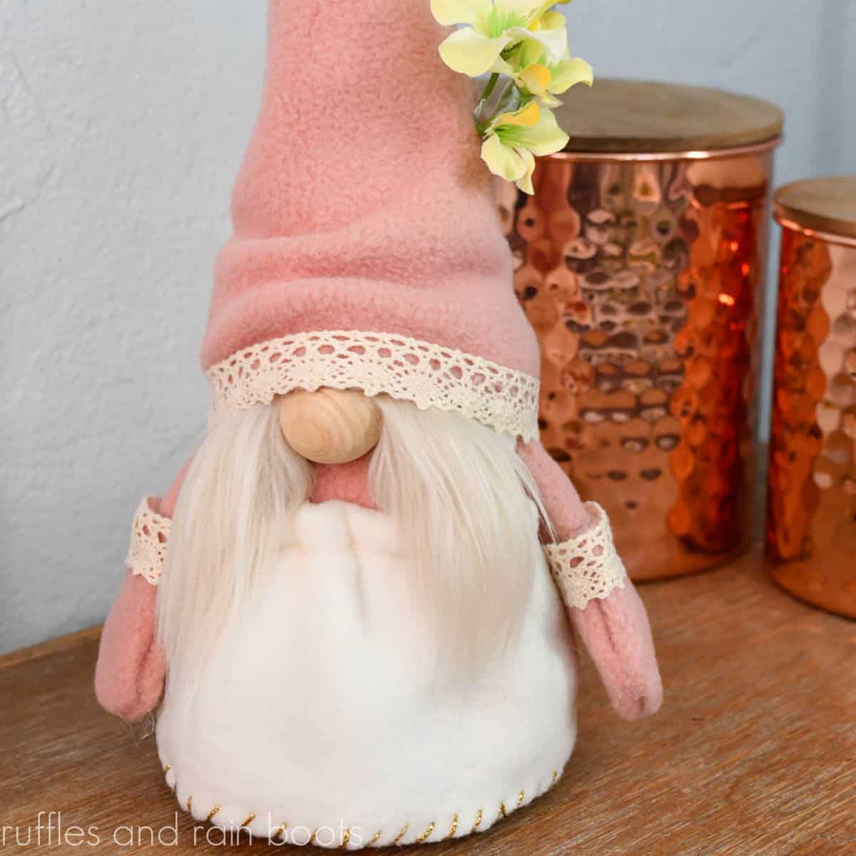 Square close up image of a girl gnome in a dress with lace and floral accents in front of copper tins on a wood table.