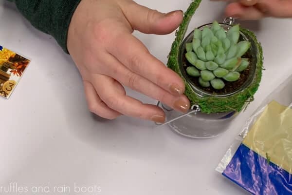 Hands gluing strips of sheet moss to the top of a hanging mason jar planter.