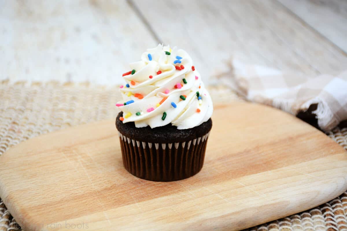 Horizontal image showing a chocolate cupcake on wood cutting board with a mound of vanilla frosting with sprinkles.