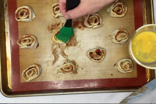Horizontal image showing a baker adding egg wash to the pastry using a green tipped silicone brush.